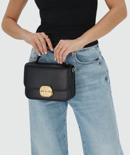 Image 9: Het model houdt een tas in het zwart met een goudkleurige bucklesluiting vast aan het handhengsel