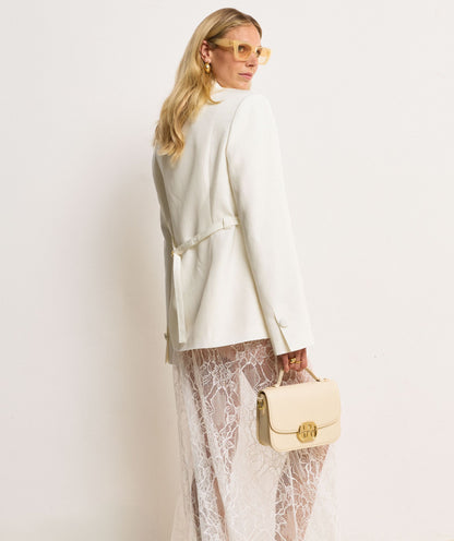 Image 13: Het model houdt een tas in Crème White met een goudkleurige bucklesluiting vast aan het handhengsel