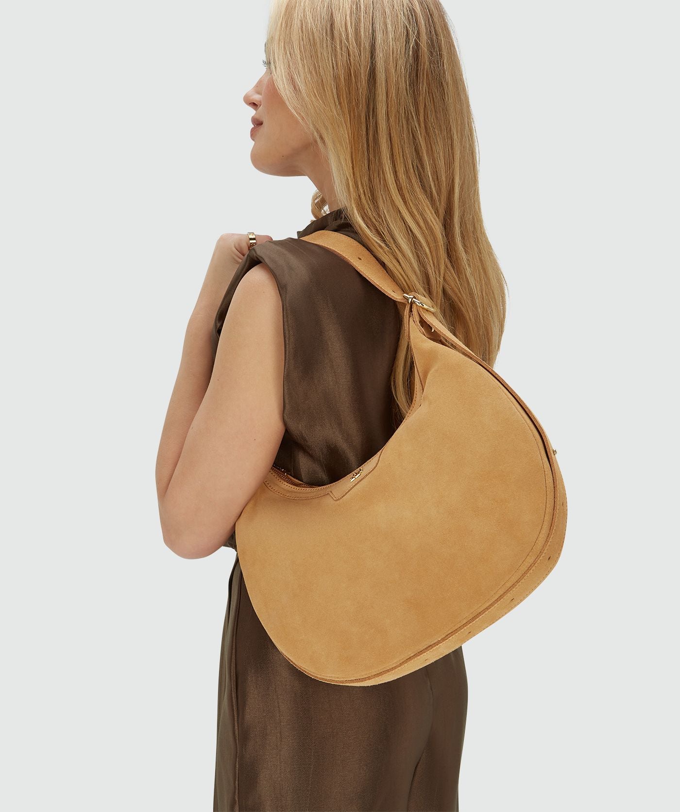 Image 22: Het model draagt een suède tas in de kleur Light Camel verkort over haar linker schouder, voorzien van een ritssluiting en een verstelbaar hengsel